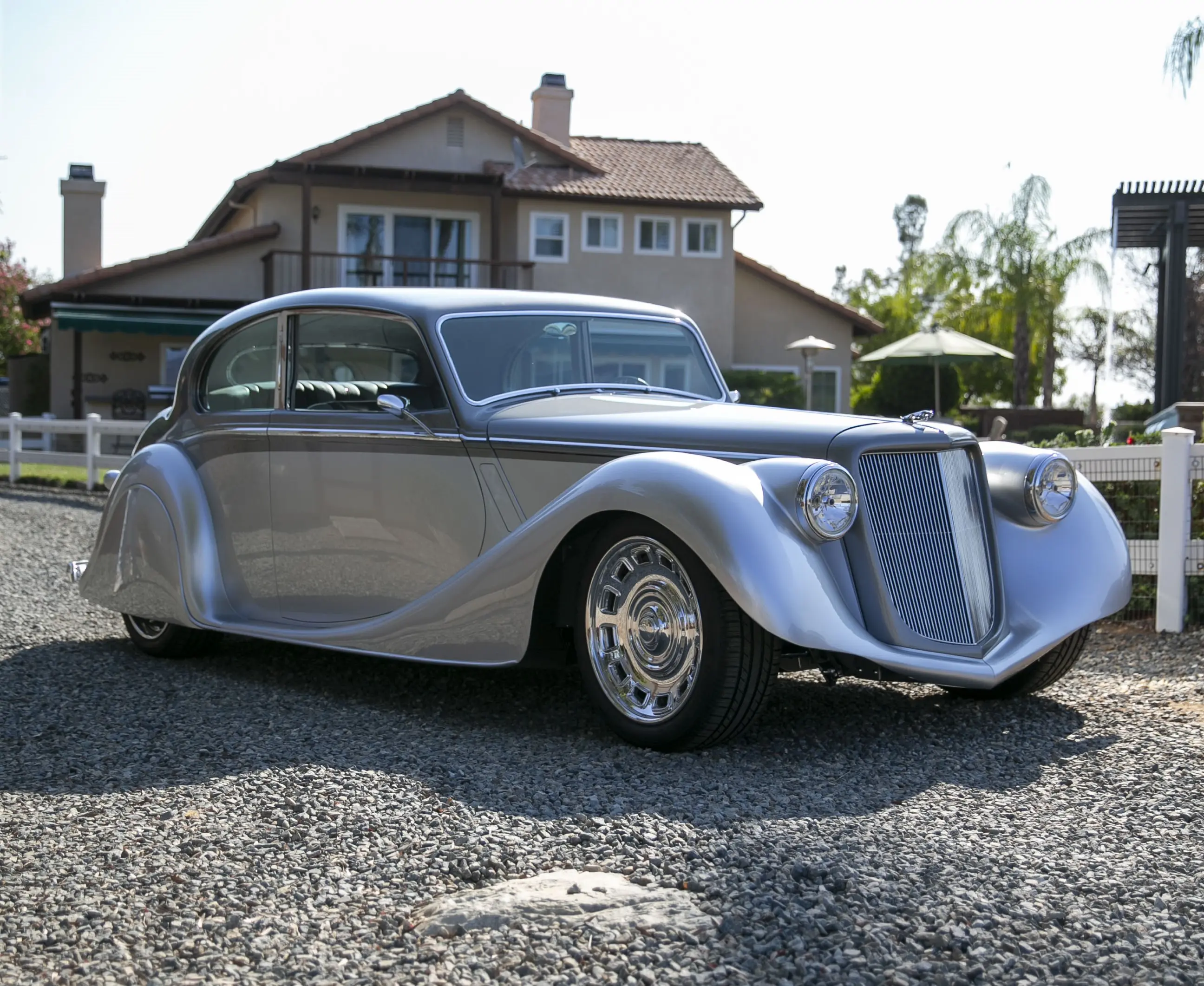 Classic silver vintage car parked on gravel in front of a house.
