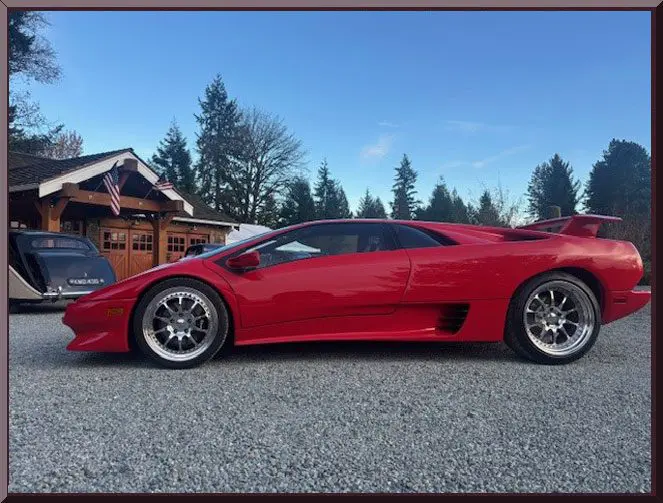 A sleek red sports car parked on gravel near a cabin with trees in the background.